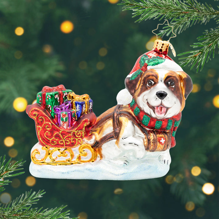 North Pole Pawtrol. Ornament of a brown and white St. Bernard pulling a red and old detailed sleigh full of presents in colors of pink, green and blue. View of front.