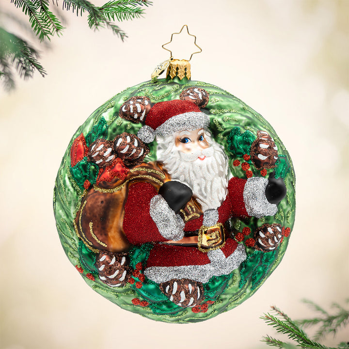 Wreathful Wishes. A round sparkling green ornament of Santa in his red suit carrying a brown bag.