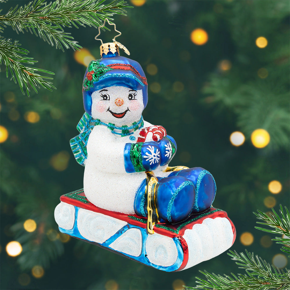 Sledding Into Cheer. Ornament of a snowman wearing matching blue gloves, hat, and boots on a red and blue sled. View of front