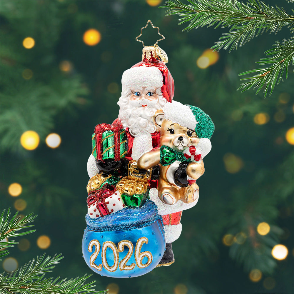 St. Nick’s 2026 Treasures. Santa carrying a teddy bear and a blue bag filled with presents and the numbers 2026. View of front.