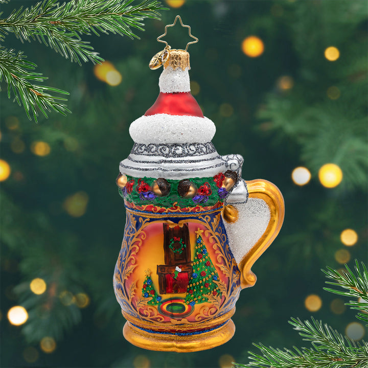 Jingle and Stein. Ornament of a golden beer mug with colors of blue, green, and a warm, cozy living room painted in the middle, and a red top hat. View of front.