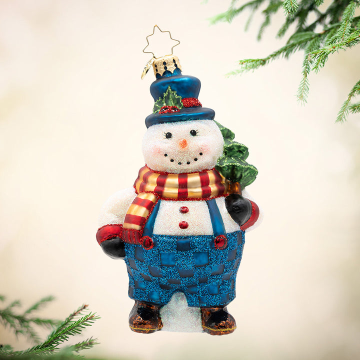 Overalls & Evergreen. A snowman ornament wearing a blue hat and sparkly overalls along with a gold and red scarf, while holding a tree. View of front