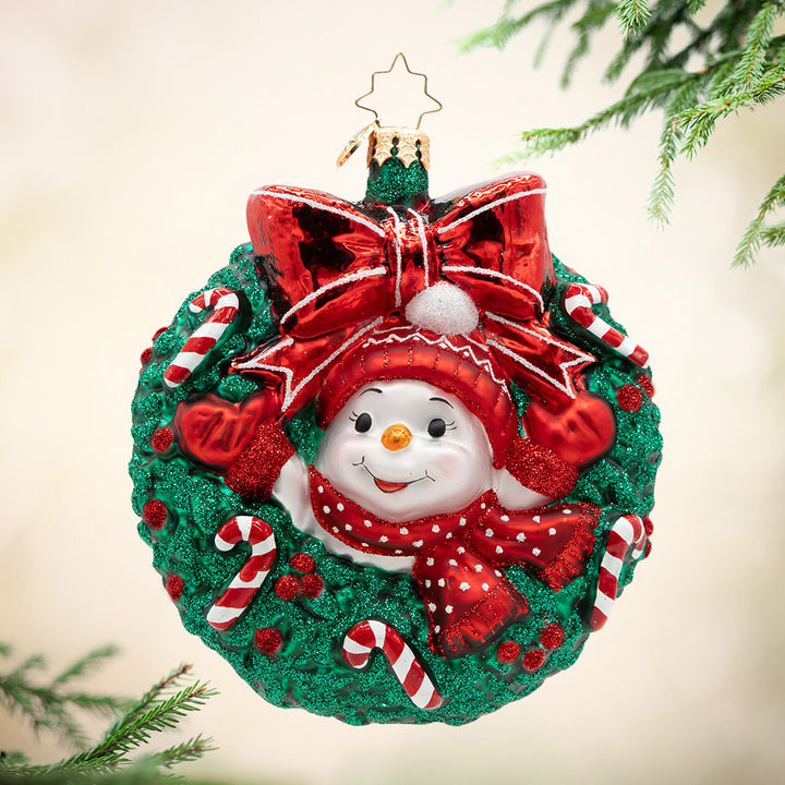 Candy Cane Cuddles. Ornament. A snowman wearing matching red hat, scarf, and mittens, popping out of a green wreath decorated in candy canes. View of front.