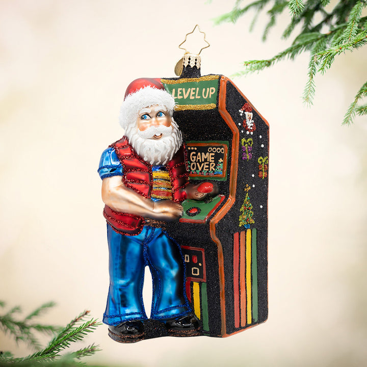 Level Up Santa. Ornament of Santa in blue pants and a red vest playing a video game machine in the color black and rainbow stripes, along with the phrase "Game Over." View of front