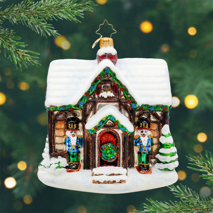 Home for the Holly-days. An ornament of a snow-covered home, complete with festive lights, a decorated tree, and two friendly nutcrackers standing guard. View of front.