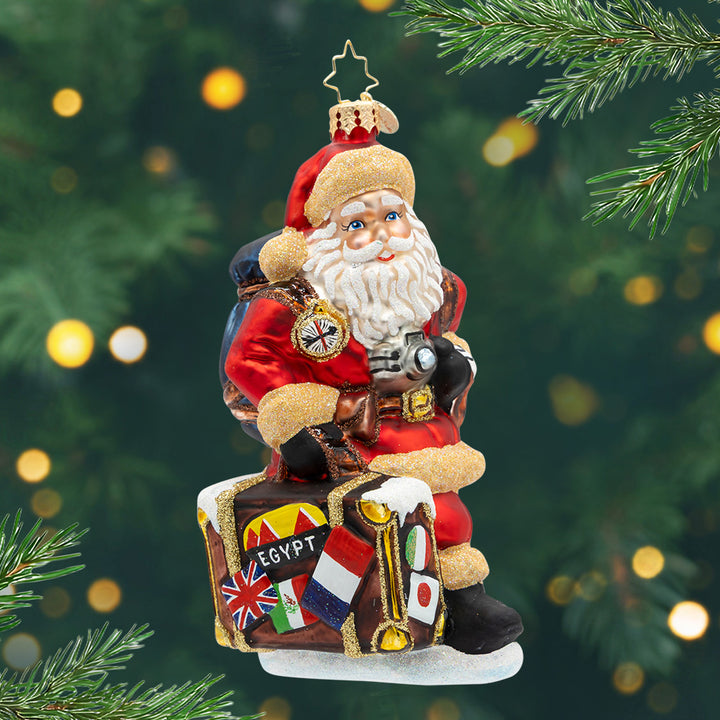Claus Around the World. Ornament of Santa traveling with a brown luggage, a blue bag, while holding a silver camera. View of front.