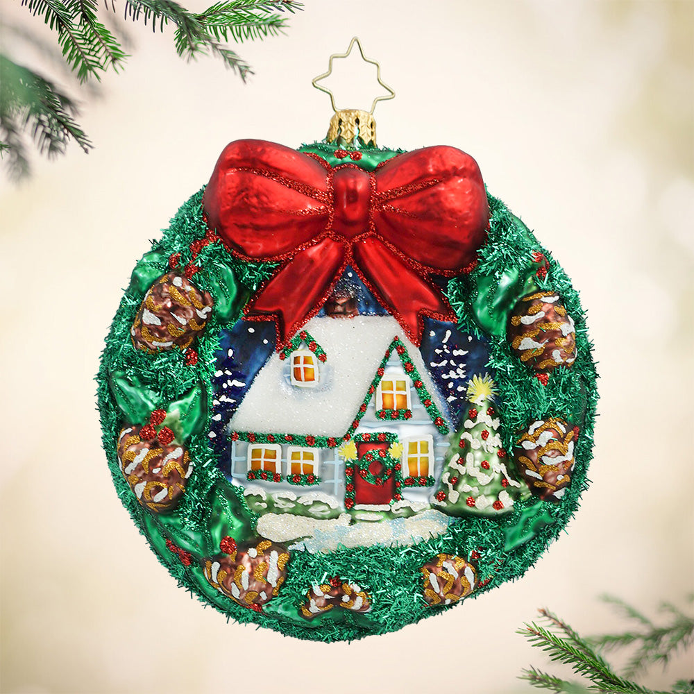 Holly Jolly Hideaway. Ornament with a night scene of a white cottage within a wreath of pinecones topped with a red bow. View of front.
