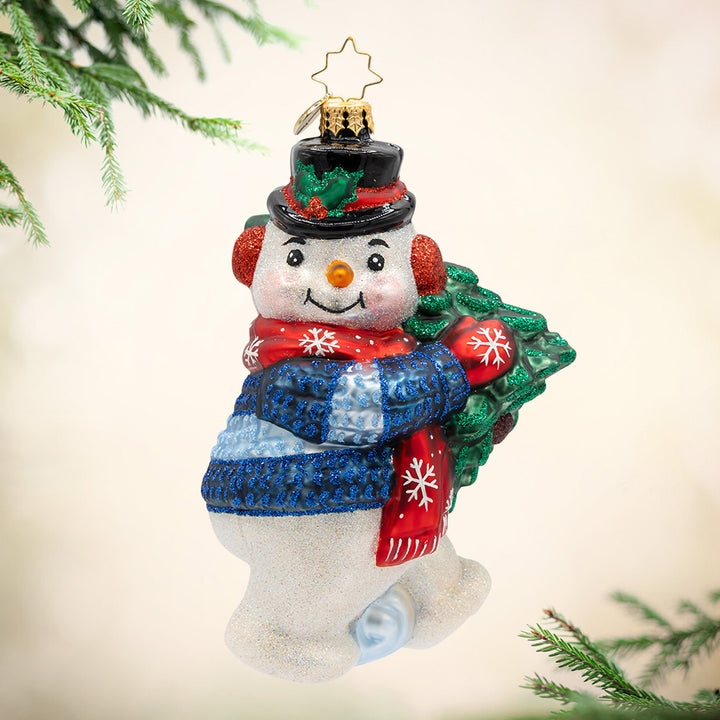 Dashing Through the Snowman. A snowman ornament wearing a blue coat  and carrying a green Christmas tree. View of front