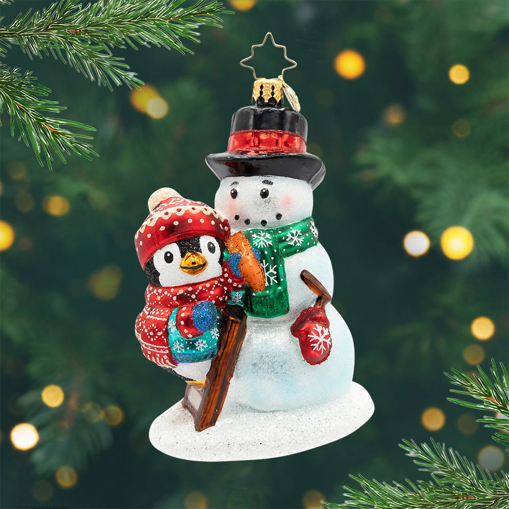 A Flurry of Friendship. Ornament of penguin wearing a blue coat and a matching red hat and scarf, finishing decorating the snowman it built. View of front