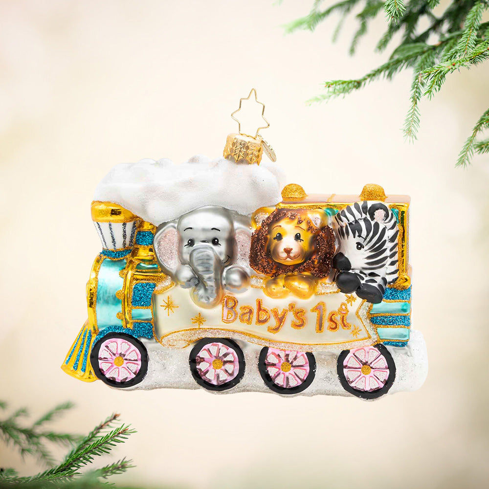 Baby's First - Tiny Travelers. A blue and gold train ornament carrying an elephant, a lion, and a zebra while holding a banner with the phrase "Baby's 1st". View of front.