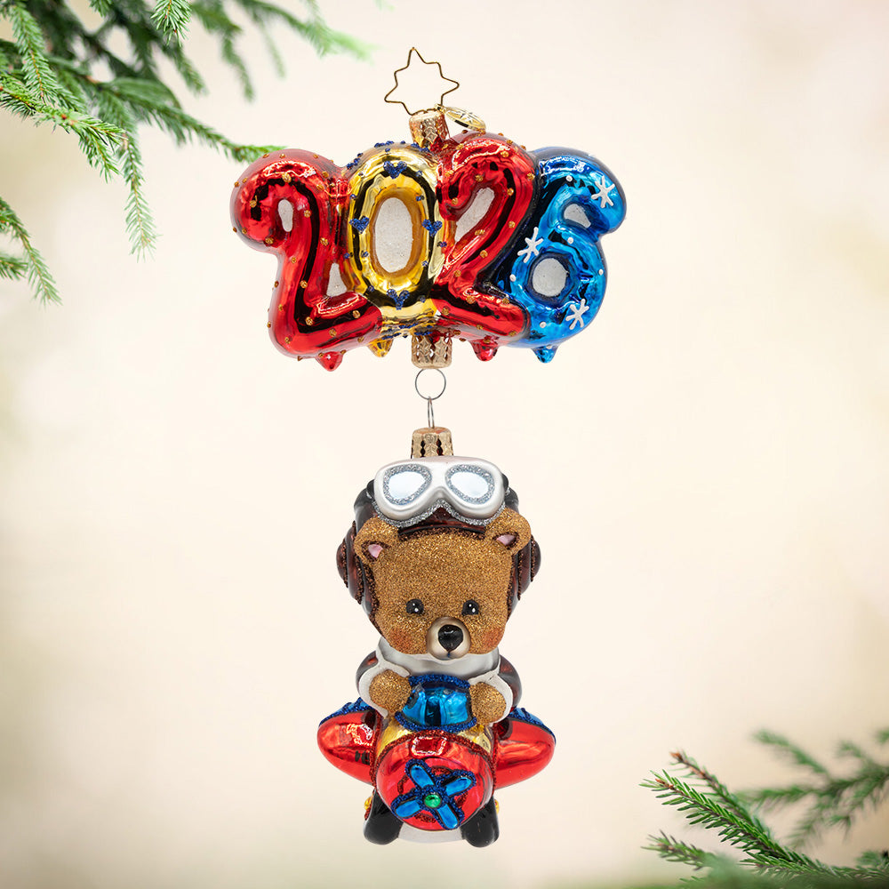 Soaring into 2026. A brown bear ornament flying on a plane that is being lifted by the numbers of "2026"  in bright red, gold. and blue. View of front.