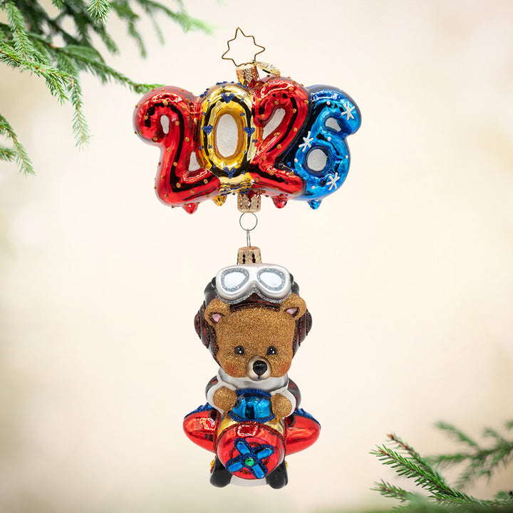 Soaring into 2026. A brown bear ornament flying on a plane that is being lifted by the numbers of "2026"  in bright red, gold. and blue. View of front.