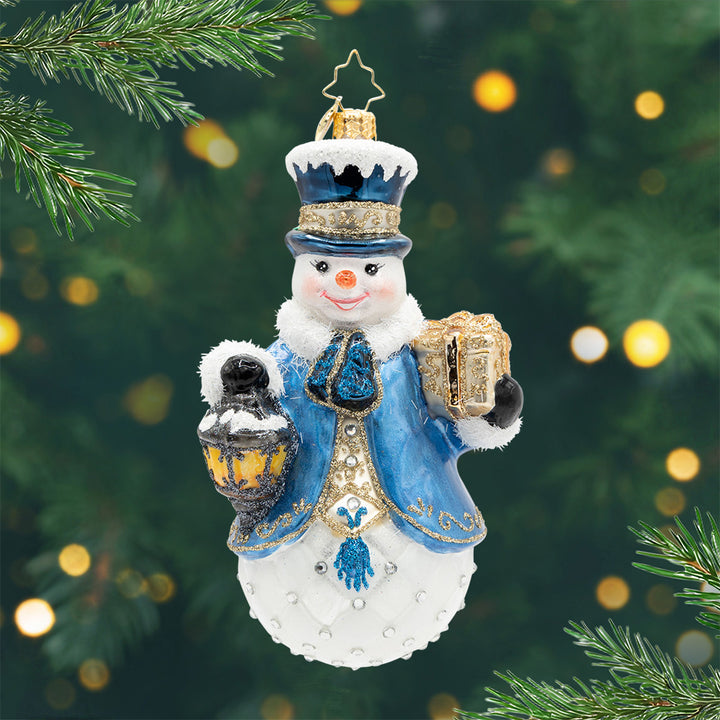 Winter Tidings. A snowman ornament with a blue coat and top hat, holding a lantern and gold present. View of front.