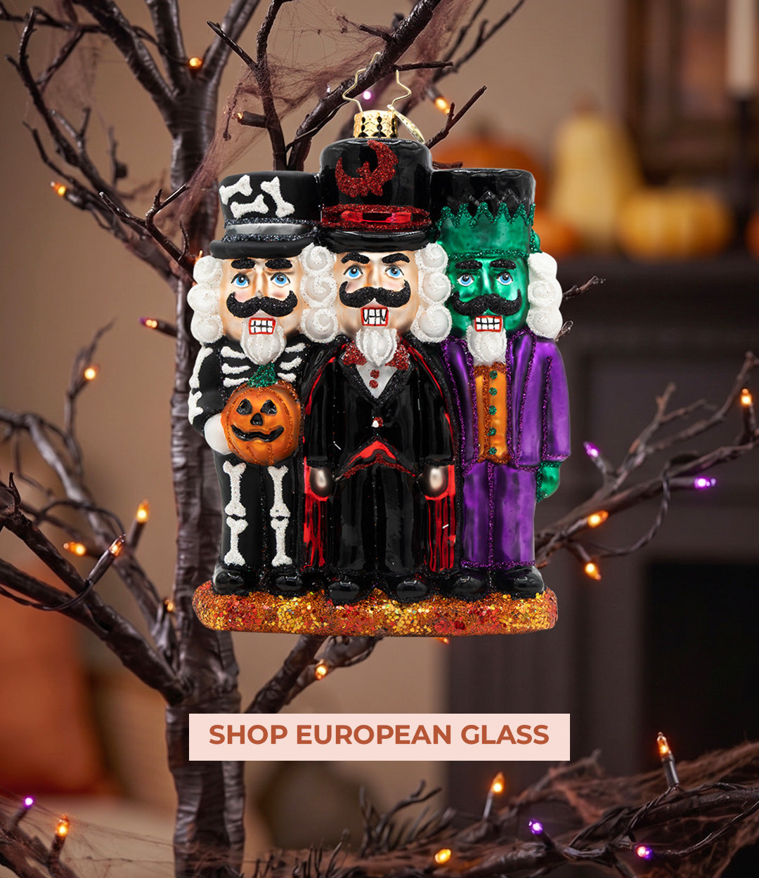 A glass ornament featuring three Halloween figures dressed as a skeleton, Dracula, and Frankenstein's monster, displayed on a spooky tree with string lights.