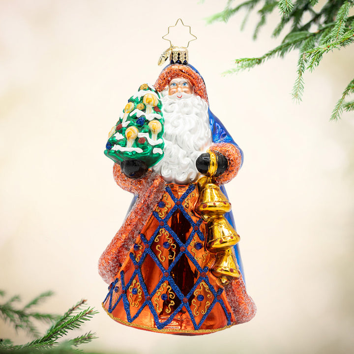 Royal Tidings Santa. This ornament of Santa dressed in a magnificent blue and copper coat, he holds a decorated Christmas tree and golden bells. View of front.