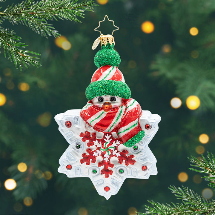 Snowflake Sparkle. A snowman ornament, wearing a glittery red, white, and green striped hat with a matching scarf, sits atop a snowflake adorned with shiny red and green gems and peppermint at its center. View of front.