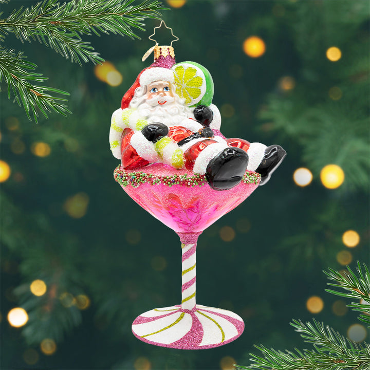 Santa's Christmas Cosmo. Ornament of Santa laying on top of a pink martini drink with a base of a pink peppermint, while holding a lime. View of front