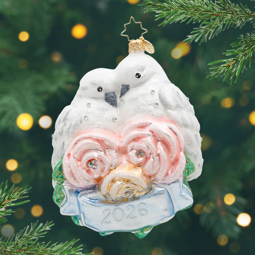 2026 Wedding Lovebirds. An ornament of two white and silver birds with three pink flowers and a blue ribbon with 2026 in front of them. View of front.