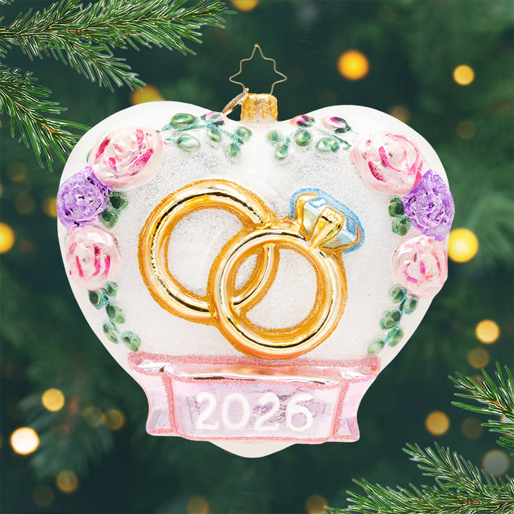 2026 Everlasting Love. A white heart shaped ornament outlined with pink and purple flowers and a pink 2026 ribbon beneath two gold wedding rings. View of front.