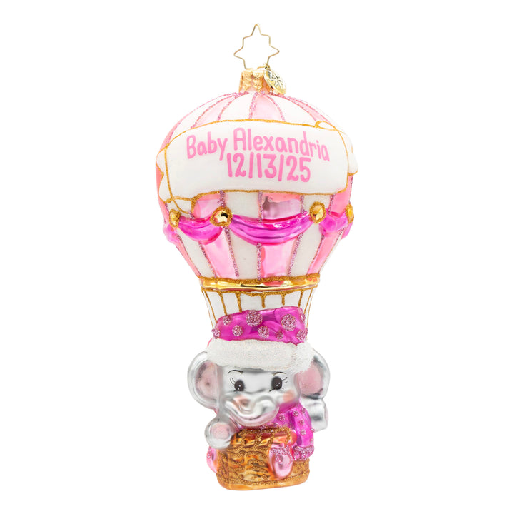 A personalized baby Christmas ornament shaped like a hot air balloon with a gray elephant in a basket. The balloon is white and pink with gold trim and a banner reading 'Baby Alexandra 12/13/25'.