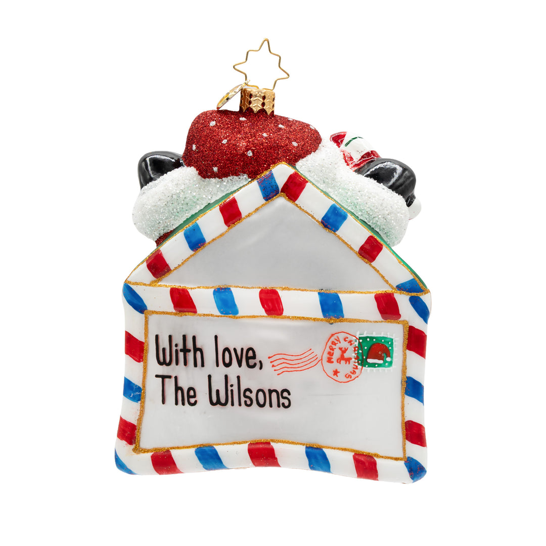 A personalized Christmas ornament shaped like a house envelope, decorated with red and blue striped borders, a red glittery hat on top, and the text 'With love, The Wilsons' printed on the front.
