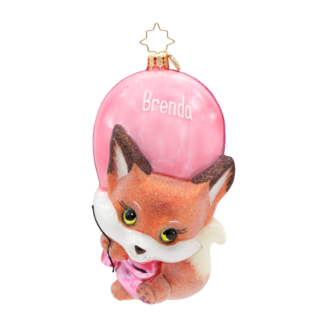 A personalized Christmas ornament featuring a cute cartoon fox wearing a pink scarf, holding a large pink balloon with the name 'Brenda' written on it, and a gold star hanging loop at the top.