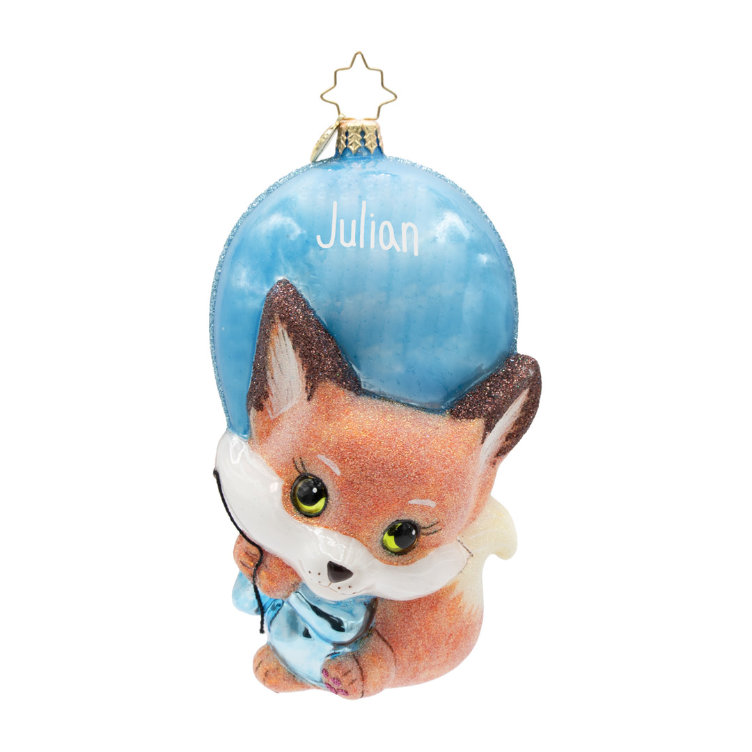 A personalized glass Christmas ornament featuring a cute cartoon fox wearing a blue outfit, with a large blue balloon behind it and the name 'Julian' written on it. The ornament is topped with a gold star hanger.