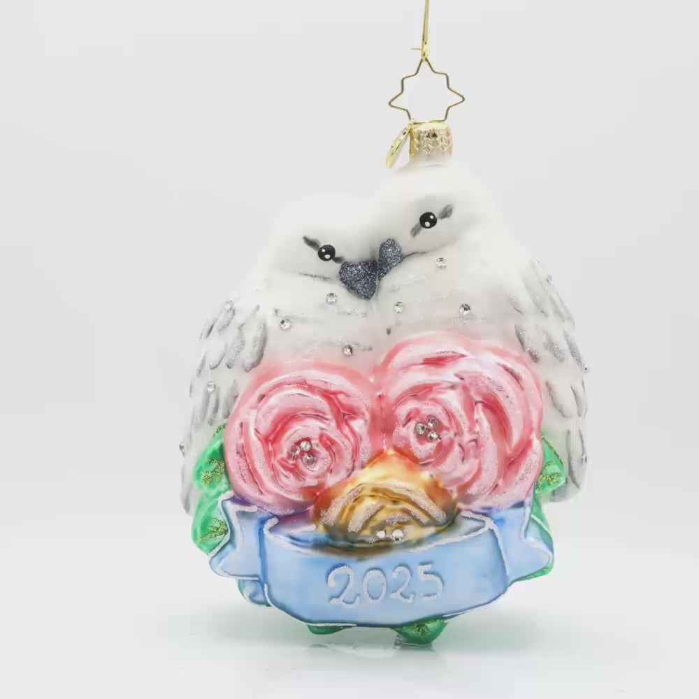 Wedding Lovebirds ornament. White birds are nestled into one another accompanied by pink flowers and a blue banner dated "2025." Video.