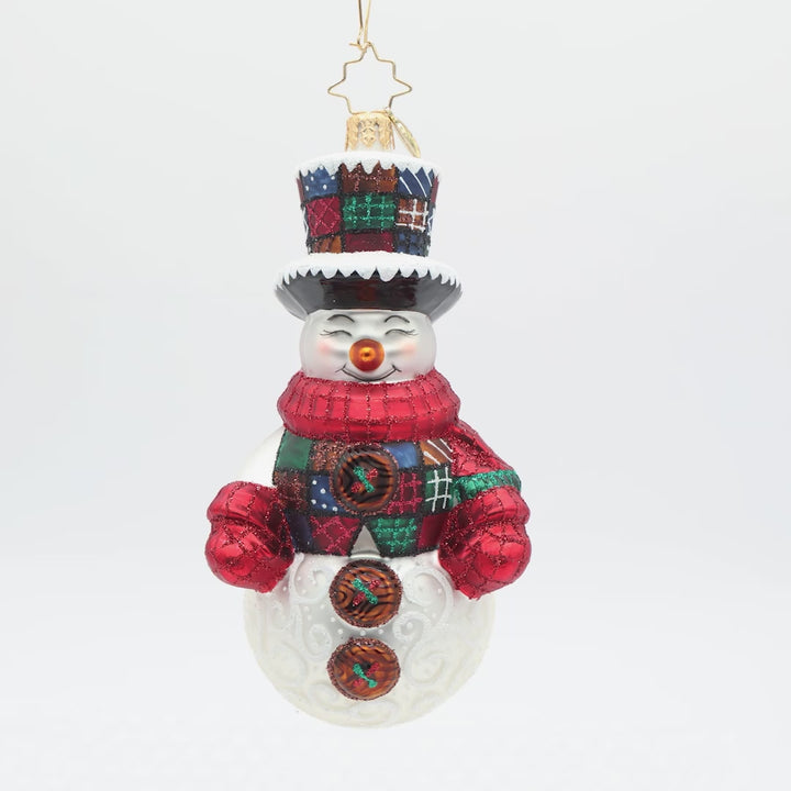Front view of a snowman Christmas ornament with a white textured body, a red glittery scarf, a black and white brimmed hat, and a colorful patchwork-patterned top hat, shown on a plain white background.