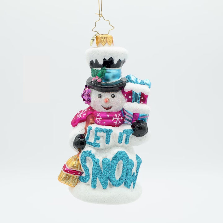 Front view of a ornament shaped like a snowman holding two presents in the colors pink and blue in one had and in the other a broom, with the phrase "Let It Snow" in blue on the front.
