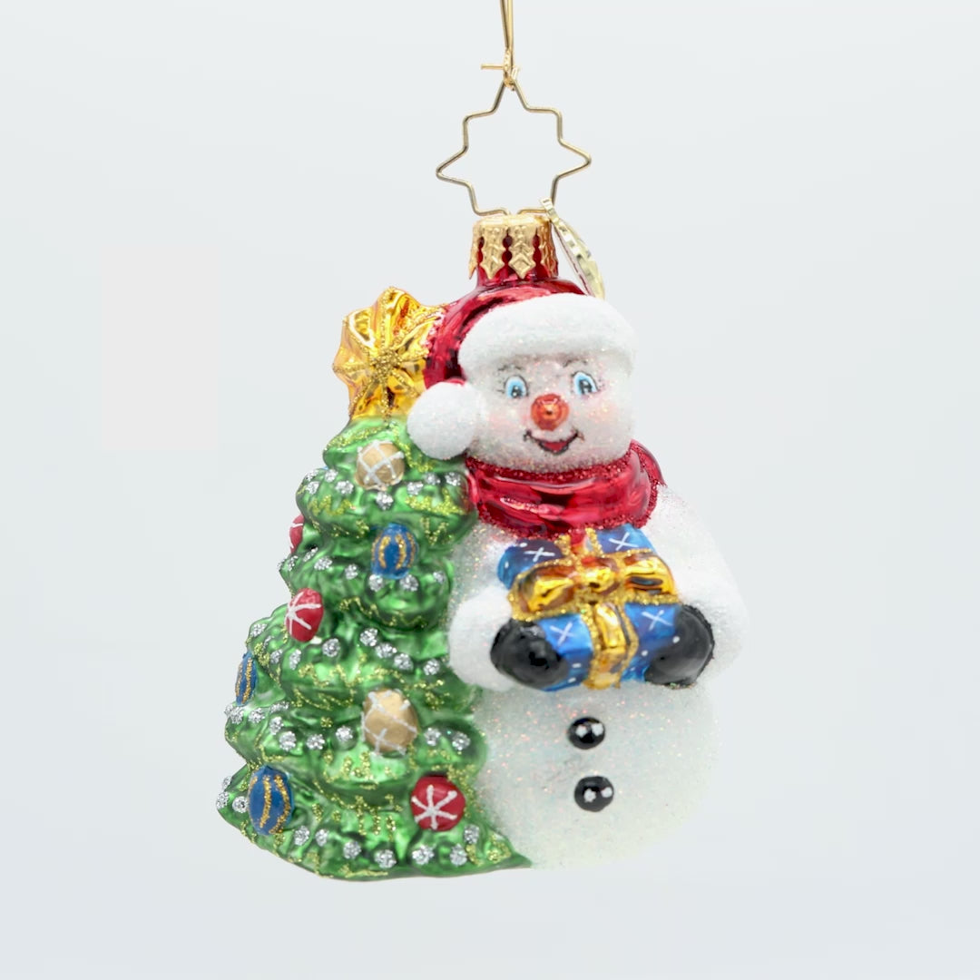 Snowy & Bright! Gem. Snowman ornament dressed in a red hat and scarf, holding a blue present next to a decorated Christmas tree. View of front