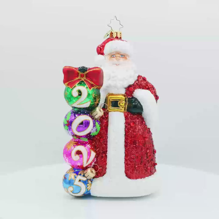 2025 Christmas Countdown ornament. Santa dons a glittering red jeweled coat.  Ornaments with "2025" written on them are stacked next to his side. Video.