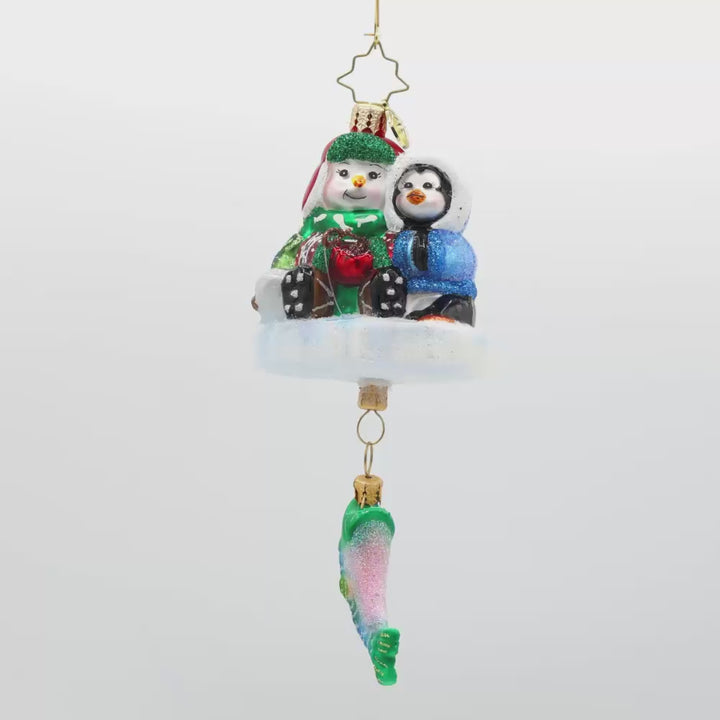 Ice Fishing Snow Pals Gem ornament. A snowman and penguin sit bundled up on a block of ice with a fishing pole. A fish dangles below. Video.