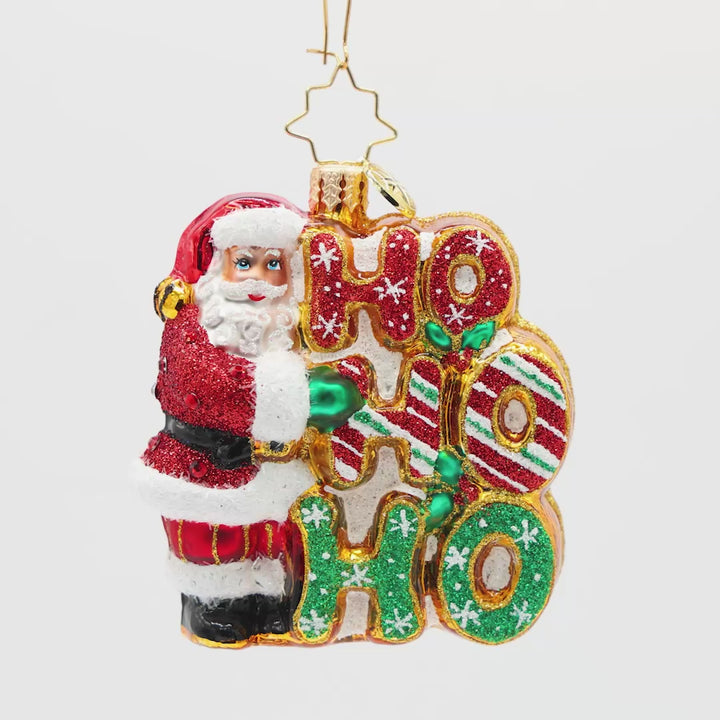 Merry Ho Ho Ho Santa Gem. Ornament of Santa in a sparkly red suit next to the phrase "HO HO HO' which are in different patterns of sparkling colors, red and green. View of front.
