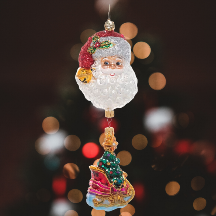 Ornament Description - Santa's Magic Sleigh: If you're looking for the perfect piece to add a unique twist to traditional Christmas imagery, this is the one! An ornate sleigh ornament, carrying a fully trimmed Christmas tree dangles from a larger ornament of Santa's smiling face.