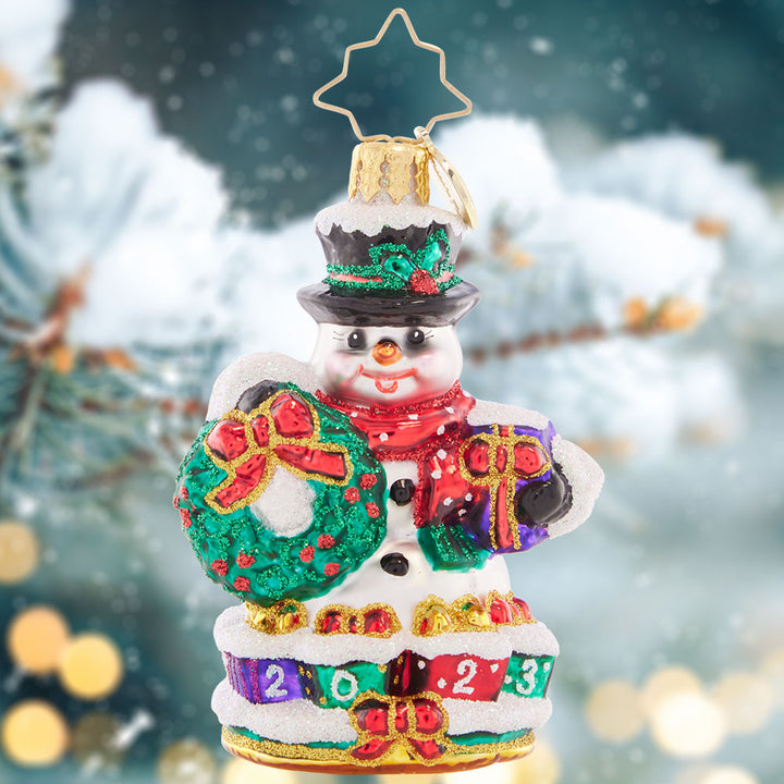 Ornament Description - Holly Jolly Snowman 2023 Gem: This jolly snowman is joyfully looking ahead to the new year. Bearing a Christmas wreath and a gilded gift, he's bringing good tidings all holiday season long.
